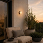 Clérachic outdoor wall light glowing on a terrace wall above an olive tree and a cushioned armchair at sunset.