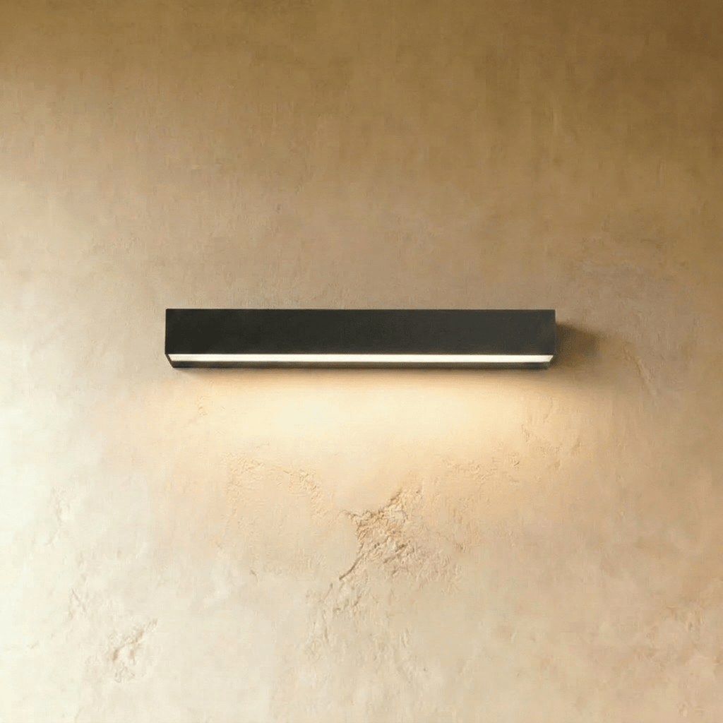 Clérachic longer slim black wall light with a lit diffuser strip glowing warm on a limewashed wall.