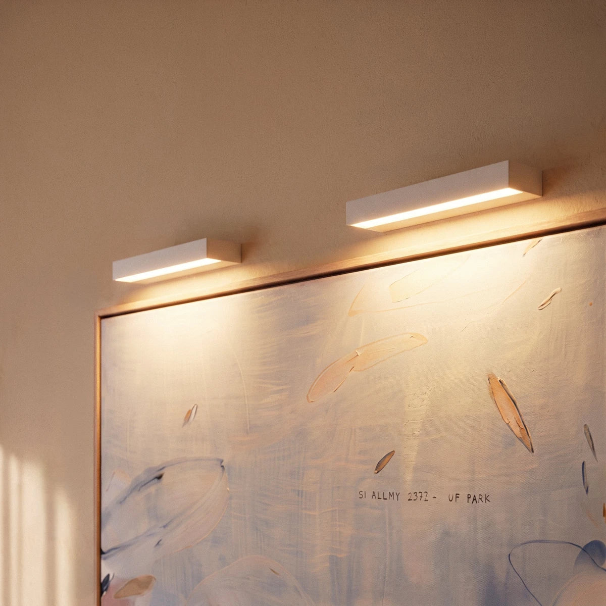 Close view of two Clérachic slim white wall lights glowing warm over the top edge of a framed painting.