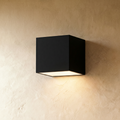 Clérachic black cube wall light mounted on cream limewash plaster, casting a soft warm downward glow from its frosted base.