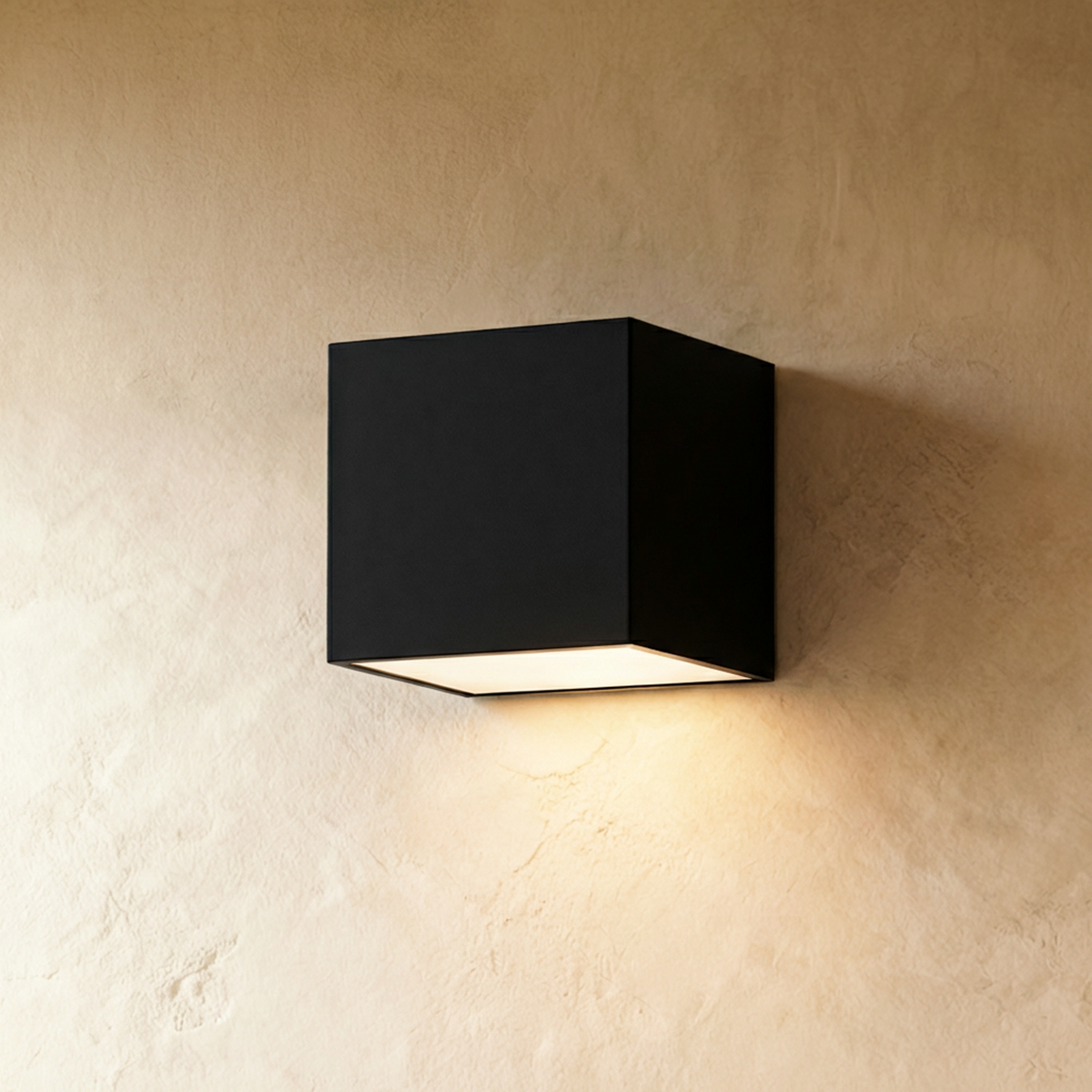 Clérachic black cube wall light mounted on cream limewash plaster, casting a soft warm downward glow from its frosted base.