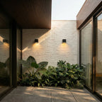 Clérachic black cube wall lights on a whitewashed brick courtyard wall, glowing warm above tropical foliage and paving.