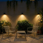 Clérachic black cube wall lights above a terrace seating area with two slatted chairs, lighting the wall and hanging vines.