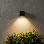 Single Clérachic black cube wall light on a rough grey plaster wall, throwing a soft downward cone over a clipped shrub.