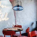 Clérachic oval blown glass pendant light in blue glass over a set restaurant table beneath a Farm to Table neon sign.