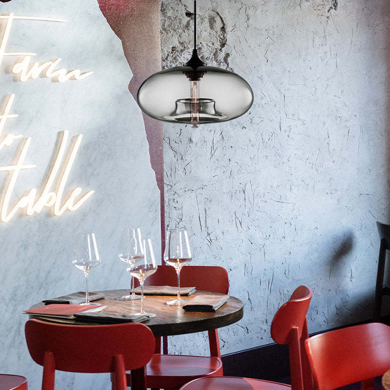 Gray glass version of the Clérachic oval pendant light hanging above a wine-set bistro table against a rough plaster wall.