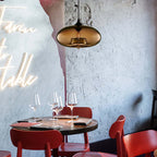 Amber glass Clérachic oval pendant light glowing over a laid restaurant table beside a marble wall and red chairs.