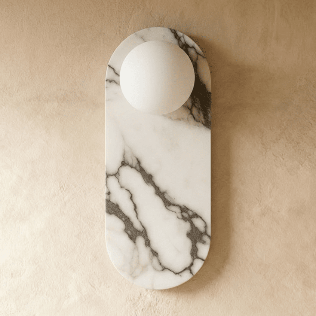 Clérachic wall light with an oval veined marble backplate and one unlit white glass globe on a plaster wall.