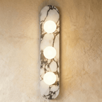Clérachic three-globe wall light on a long rounded marble backplate, all three glass globes glowing warm on a plaster wall.