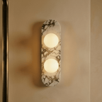 Close view of a Clérachic two-globe wall light on a veined marble backplate, glowing warm on a smooth painted wall.