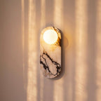Clérachic one-globe wall light on an oval veined marble backplate, warm glow on a wall striped by window light.