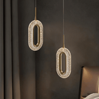 Pair of Clérachic oval pendants in brass and textured acrylic hanging at different heights against a dark wall beside a bed.