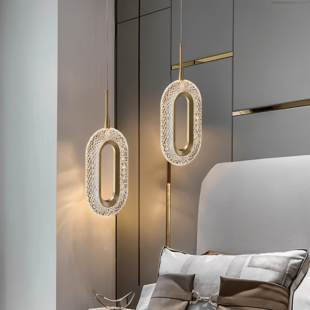 Two Clérachic oval brass and acrylic pendants lit warm beside a grey upholstered headboard and cushions in a bedroom.