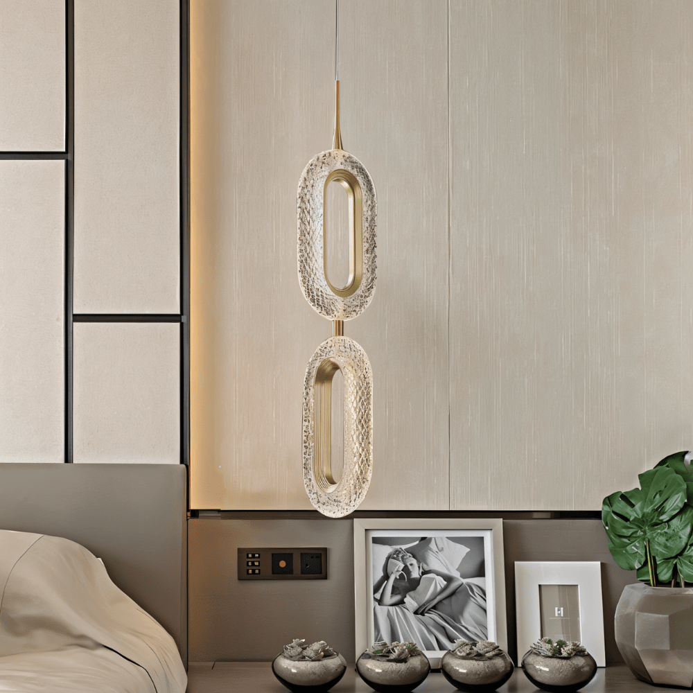 Clérachic two-head oval pendant with brass inner rings and patterned acrylic, hung beside a bed against pale panelled walls.