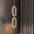 Close view of the Clérachic two-head pendant, its textured acrylic ovals and brass rings against a dark wall and heavy curtain.
