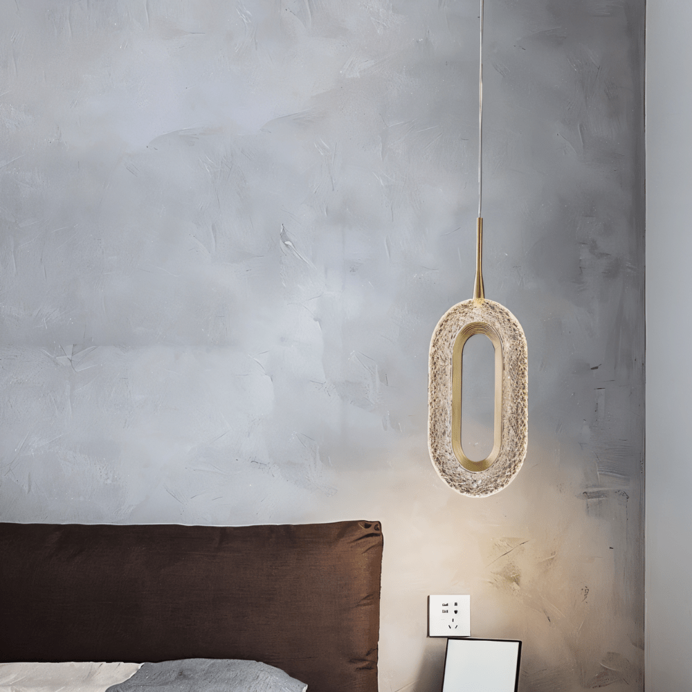 Clérachic single oval pendant in brass and textured clear acrylic hanging beside a bed against a grey limewashed wall.