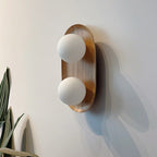 Two frosted globes in a natural wood oval frame, this Clérachic wall light throws a long shadow across a pale wall beside a leafy plant.