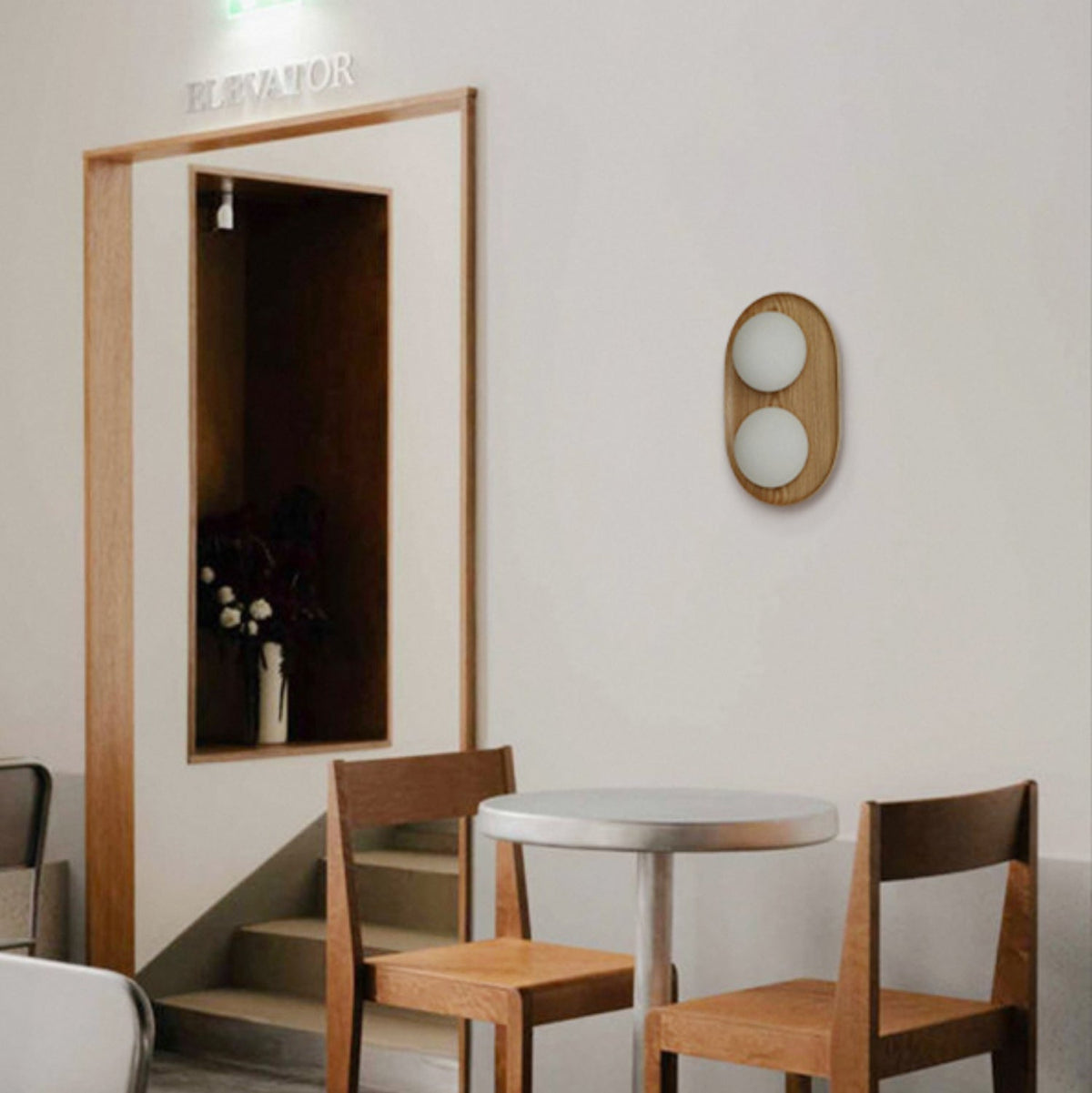 A small Clérachic wall light with two globes in a wood oval frame, hung by a door frame above a pair of wooden chairs and a round table.