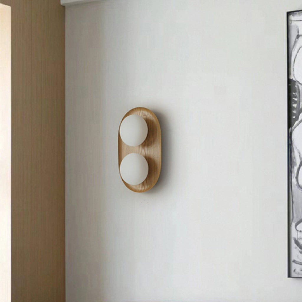 Clérachic oval wall light with two frosted globes in a pale wood frame, mounted on a plain grey wall beside a framed artwork.