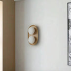 Clérachic oval wall light with two frosted globes in a pale wood frame, mounted on a plain grey wall beside a framed artwork.