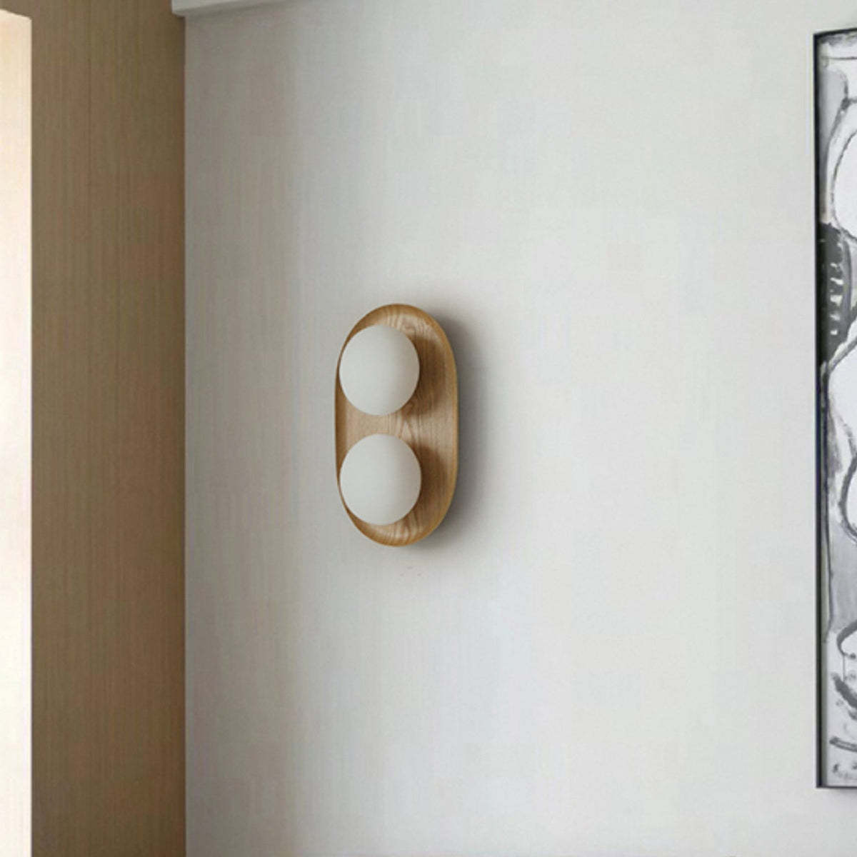 Clérachic oval wall light with two frosted globes in a pale wood frame, mounted on a plain grey wall beside a framed artwork.