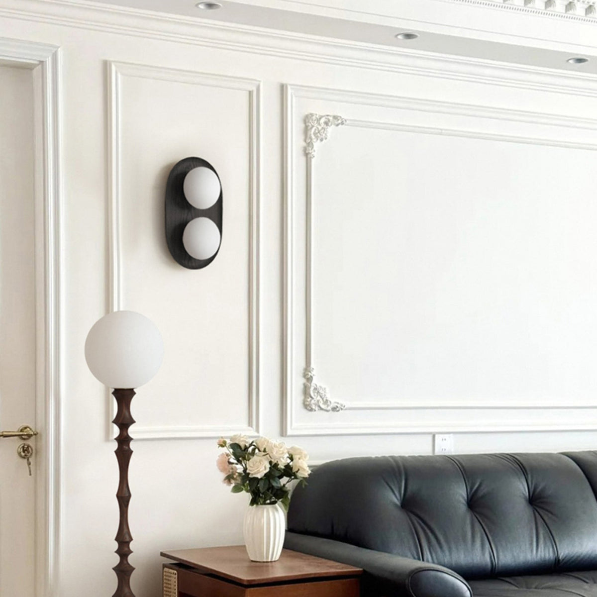 Black-framed Clérachic wall light with two frosted globes on a white panelled wall, above a leather sofa and a tall globe floor lamp.