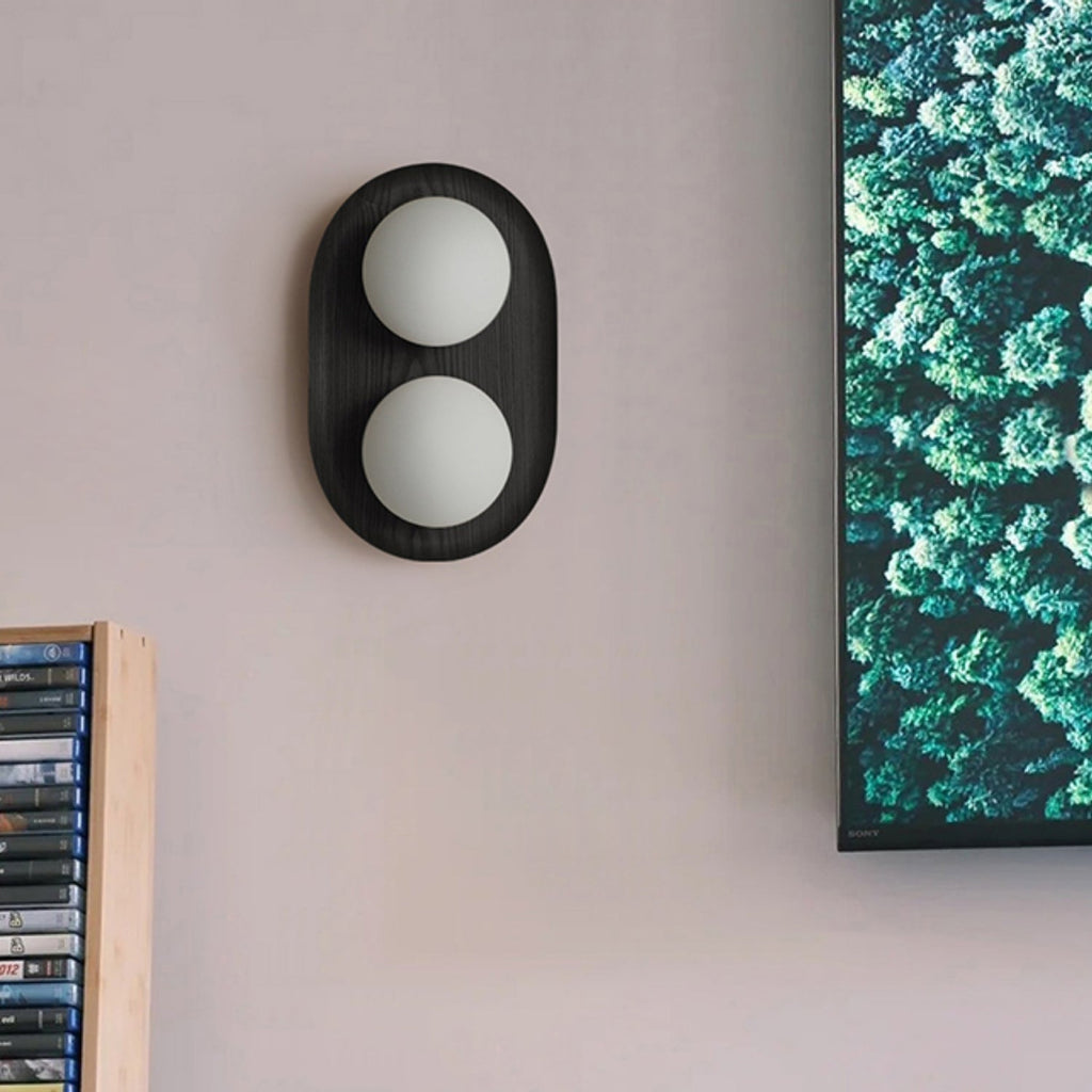 A black oval Clérachic wall light with two frosted globes on a dusky pink wall, beside a screen showing a forest photograph.