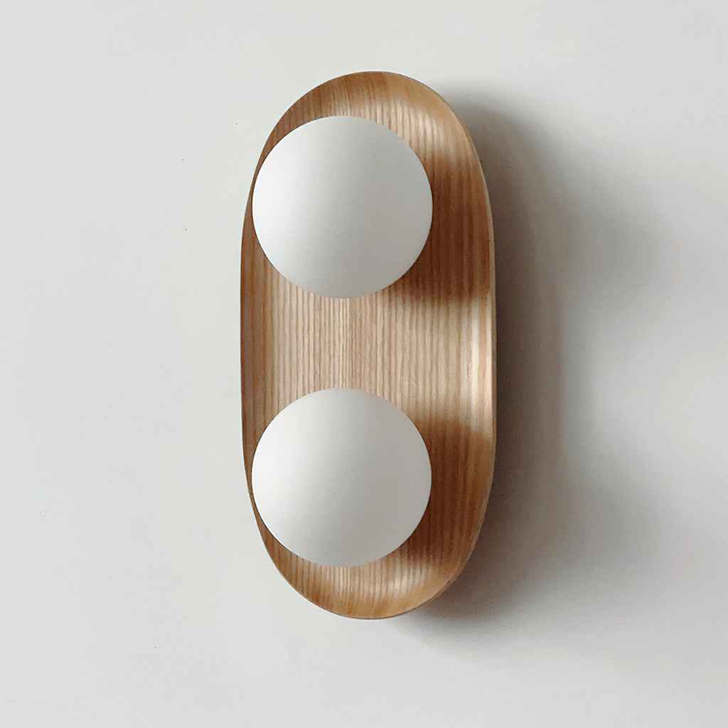 Large view of this Clérachic wall light in natural wood, its two frosted globes standing proud of the oval frame on a pale wall.