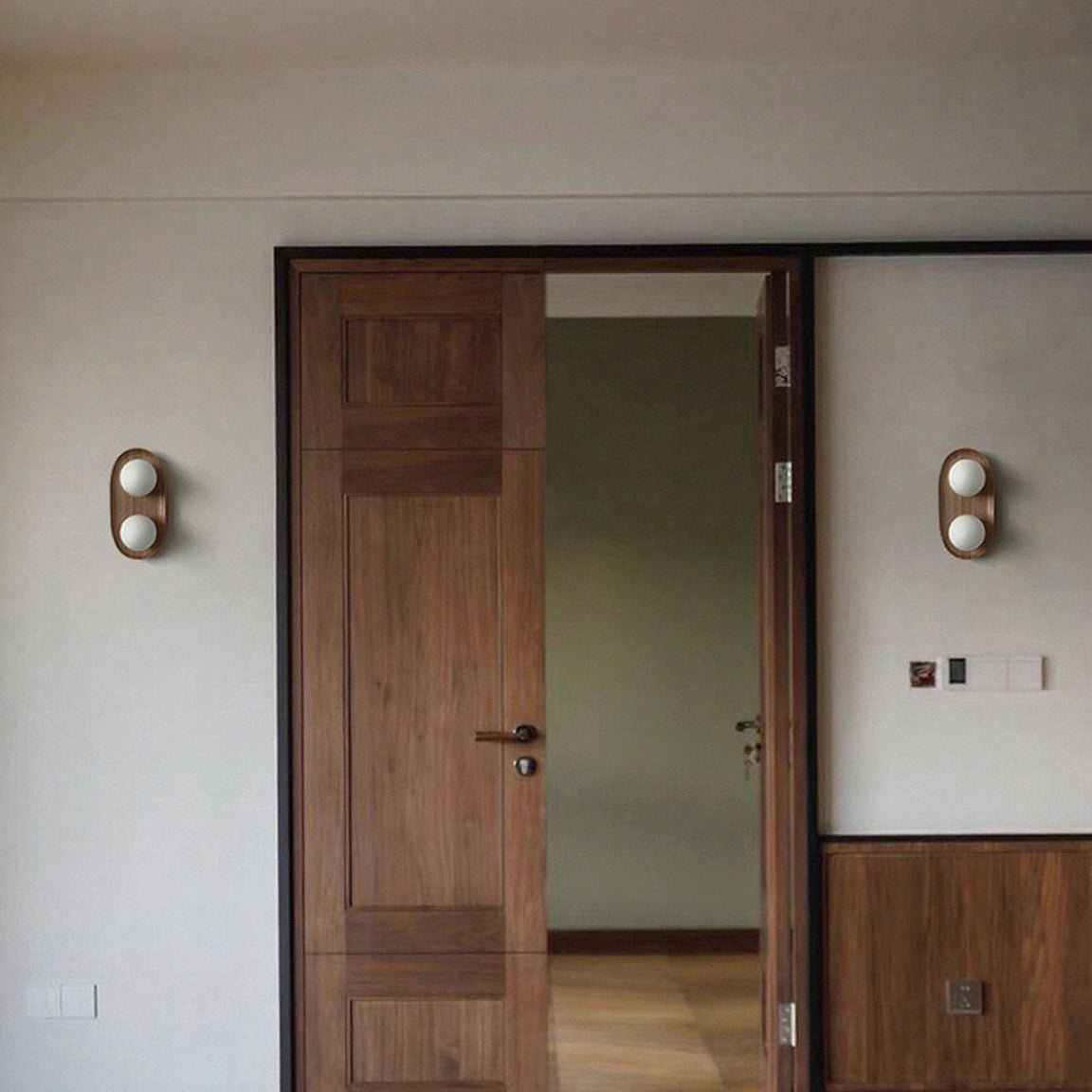 A pair of Clérachic wall lights with wooden oval frames flank a walnut double door in a plain-walled hallway.