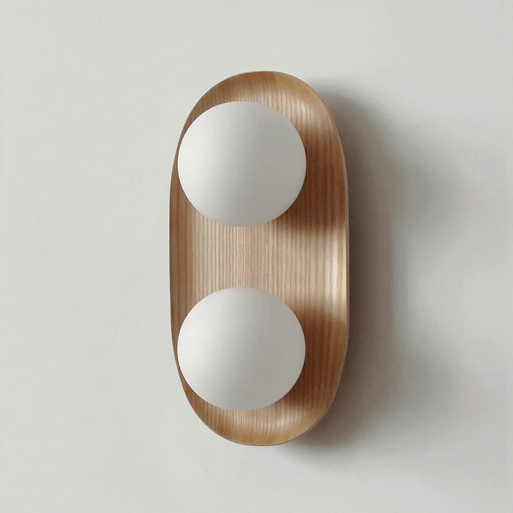 Front view of this Clérachic wall light in natural wood, two frosted globes on an oval frame, against a pale grey wall.