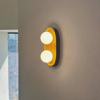 Lit Clérachic wall light with two glowing globes on a golden wood frame, against a taupe wall beside a tall window.