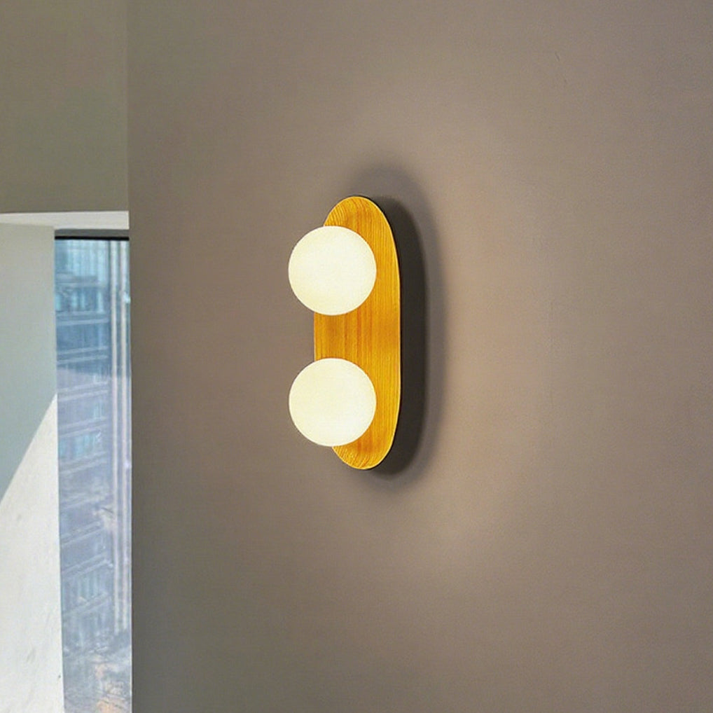Lit Clérachic wall light with two glowing globes on a golden wood frame, against a taupe wall beside a tall window.