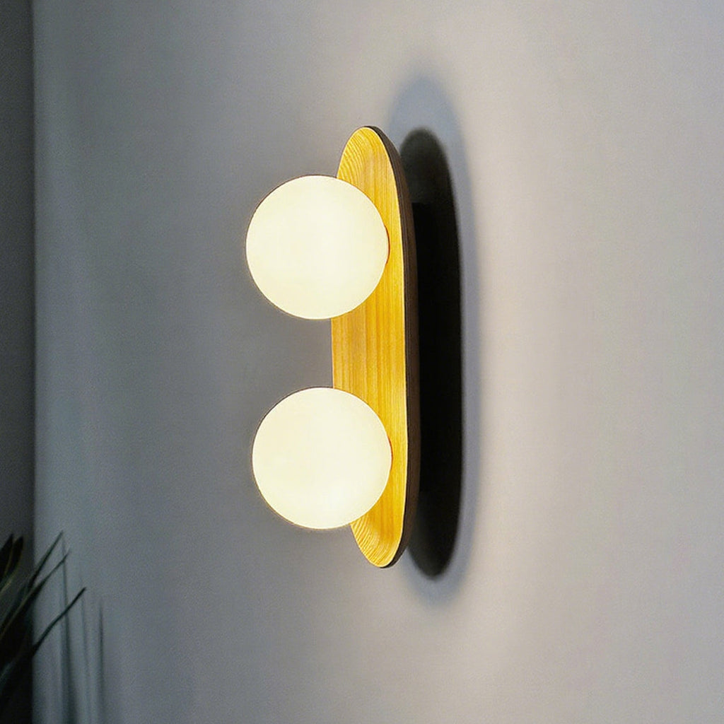 Closer view of the lit Clérachic wall light, two warm-white globes on a golden wood oval with a dark shadow on the wall.