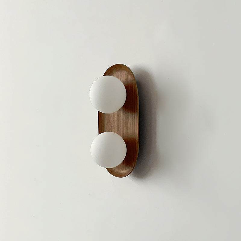 The smaller Clérachic wall light in a wood finish, two frosted globes on a slim oval frame, shown on a pale wall.