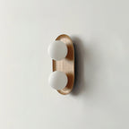 Small Clérachic wall light in a warm wood finish, two frosted globes on a slim oval frame on a light grey wall.