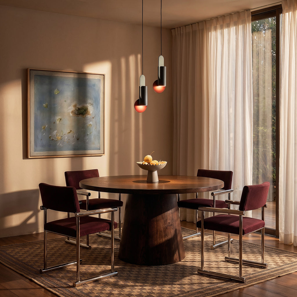 Clérachic pair of dark cylinder pendants with red-orange discs hung over a round walnut dining table by sheer curtains.
