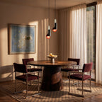 Clérachic pair of dark cylinder pendants with red-orange discs hung over a round walnut dining table by sheer curtains.
