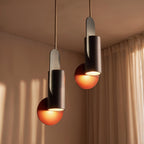 Clérachic two dark metal cylinder pendants on braided cords, their copper-red discs glowing beside sheer curtains.