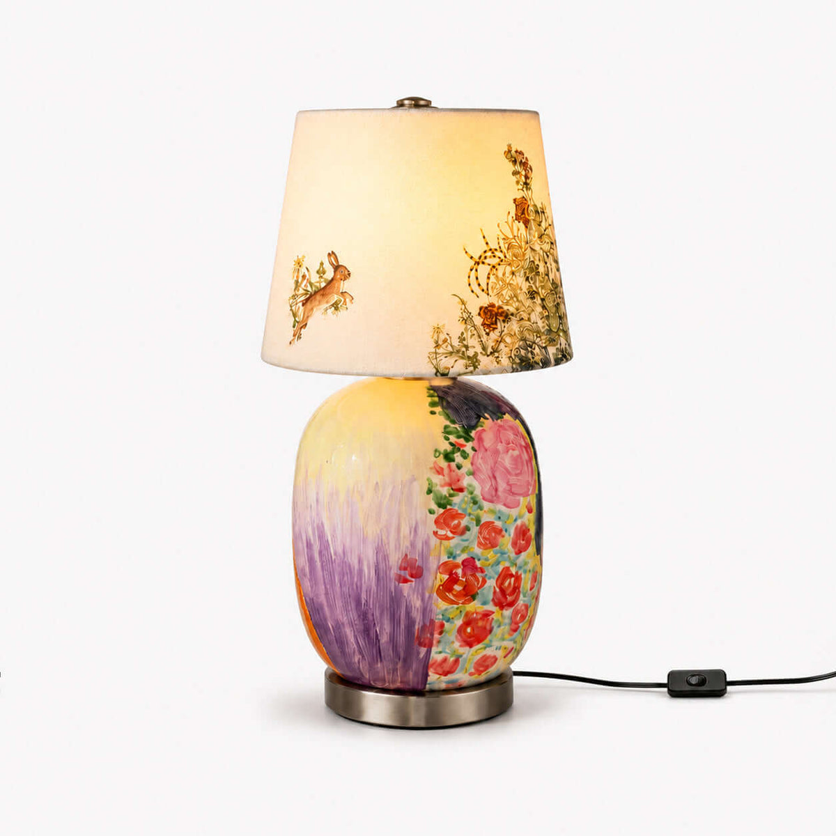 This Clérachic table lamp on a plain white backdrop, with a floral and lilac painted ceramic base and a shade printed with a hare.
