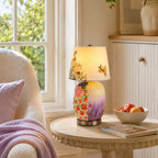 Lit Clérachic table lamp with a floral painted base on a round side table by a sunny window, next to a fruit bowl and a book.