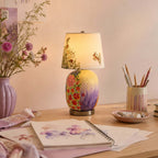 A Clérachic table lamp with a floral ceramic base on a desk, beside a vase of wild flowers, a pot of pencils and a watercolour sketchbook.