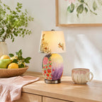 Floral-painted Clérachic table lamp on a pale wood console, with a basket of lemons and pears, a pink cloth and a patterned mug.