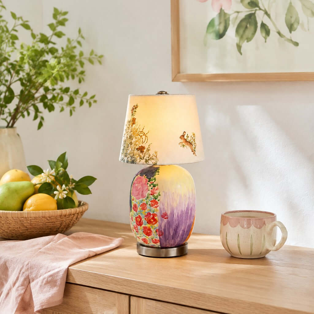 Floral-painted Clérachic table lamp on a pale wood console, with a basket of lemons and pears, a pink cloth and a patterned mug.