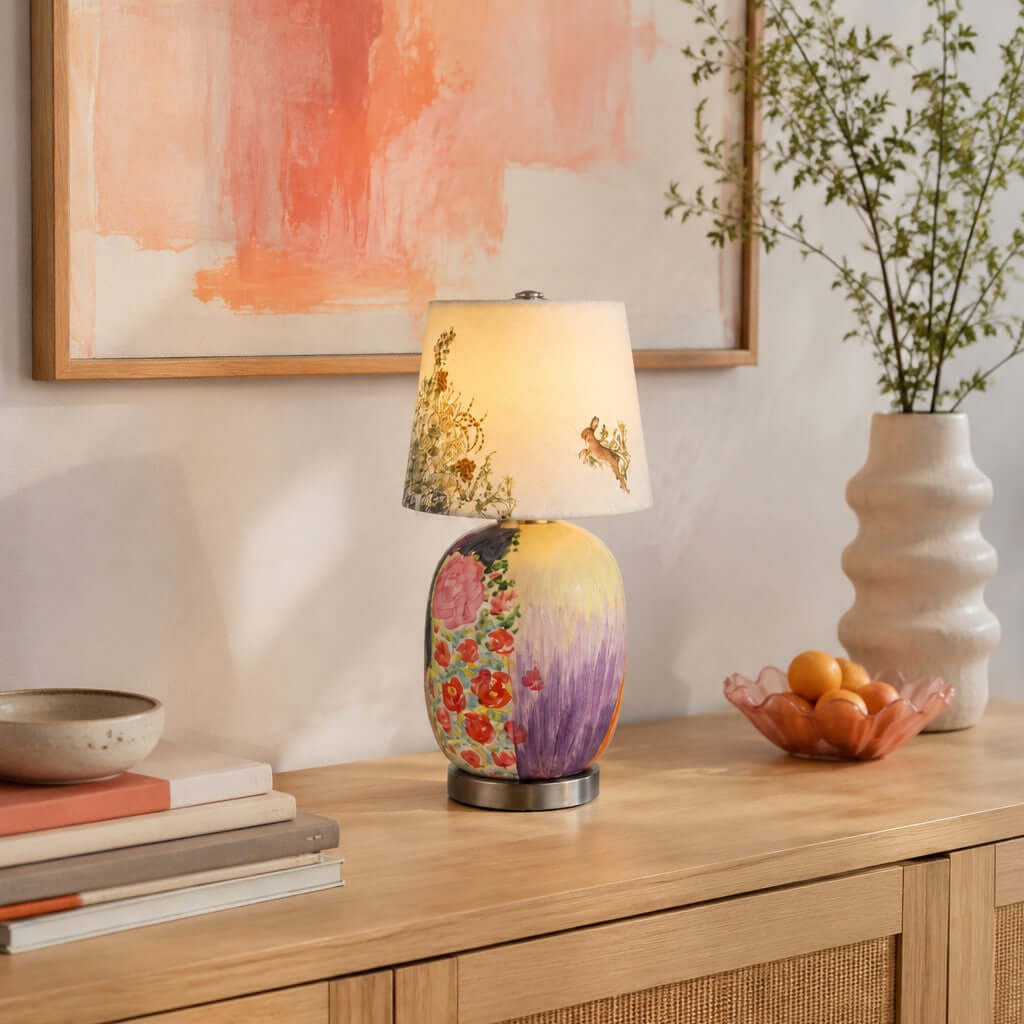 A Clérachic table lamp with a floral and lilac base on a wood sideboard, under a peach abstract print, with a bowl of oranges.