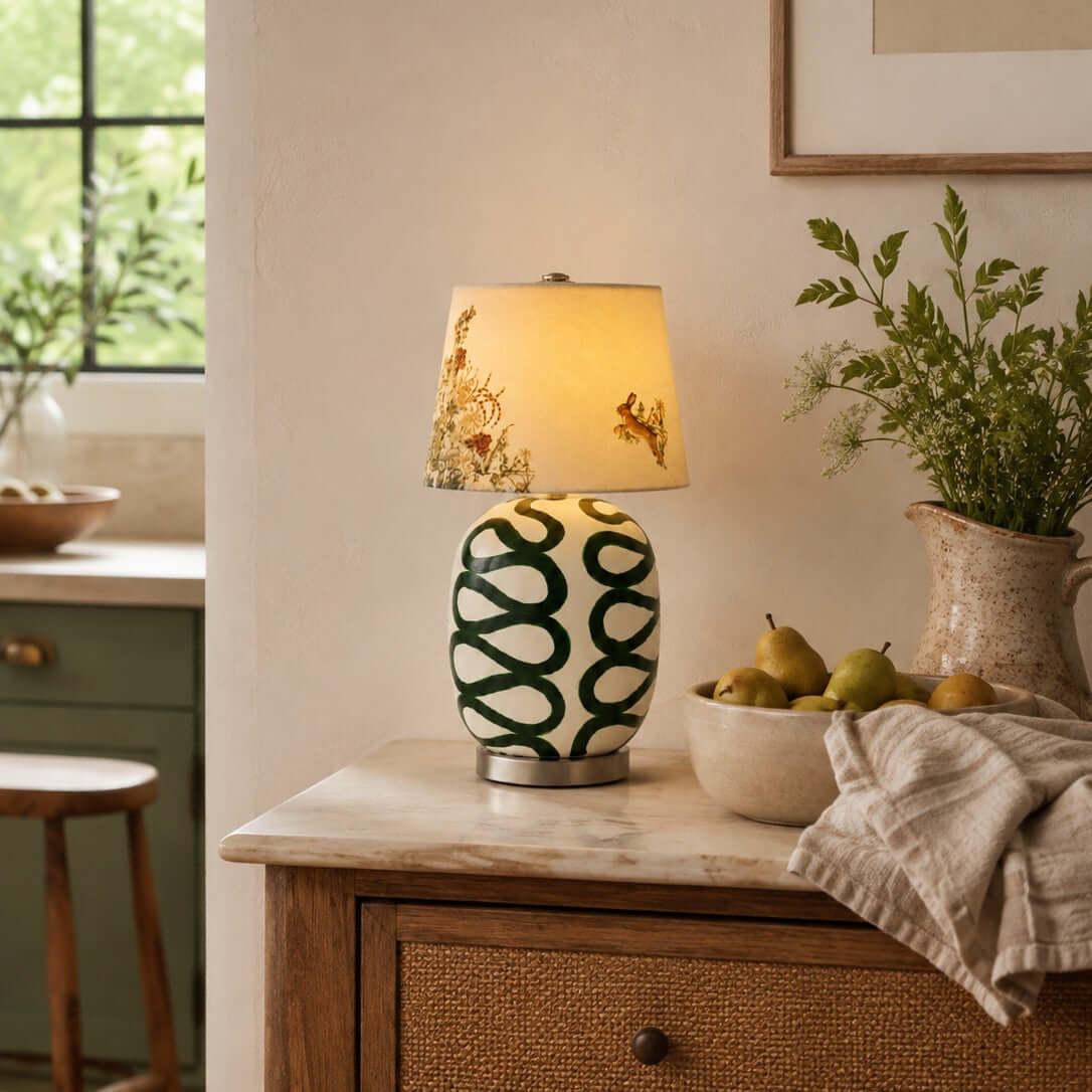 Lit Clérachic table lamp with green looped glaze on a rattan-drawered cabinet, with a bowl of pears and a linen cloth.