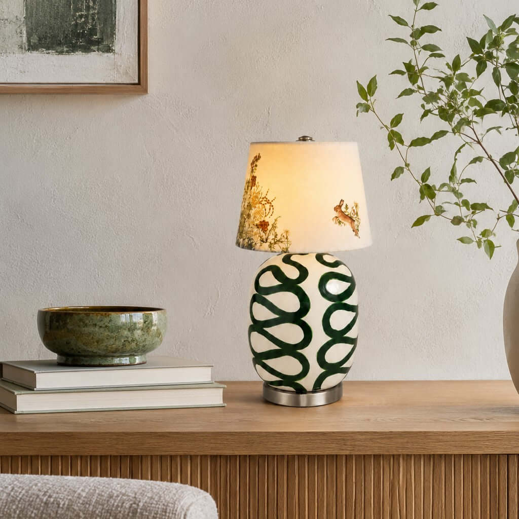 Green looped Clérachic table lamp on a wood sideboard, with a green glazed bowl, two books and a leafy branch against a white wall.