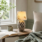 This Clérachic table lamp with black zebra-style stripes on cream sits on a curved side table by a window, with a mug and an open book.