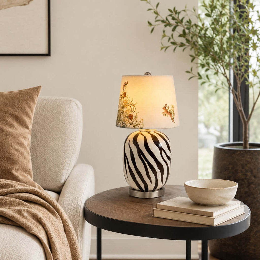 A Clérachic table lamp with a striped cream and black base on a round dark side table, beside a sofa, a stoneware bowl and books.