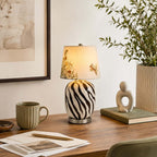 Clérachic table lamp with a striped base on a wood desk, beside a green mug, loose papers, a pen and a small sculpture.