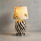 Close view of this Clérachic table lamp's striped base and shade with a hare and wild flowers, on a concrete floor.
